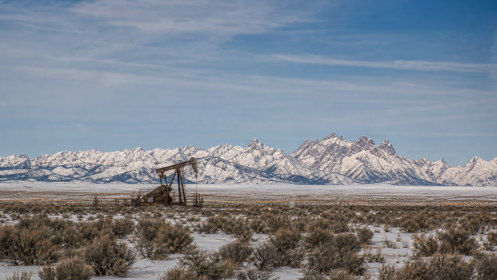 Lone weathered pumpjack on sagebrush flats with snow patches, jagged snow-covered Wind River Range across the horizon, Fremont County, Wyoming.