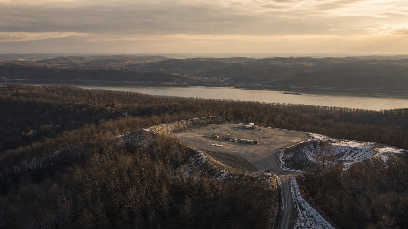 Aerial of a flattened-hilltop Marcellus pad above the upper Ohio River valley in winter, a barge tracking through the gray water below, Marshall County, West Virginia.