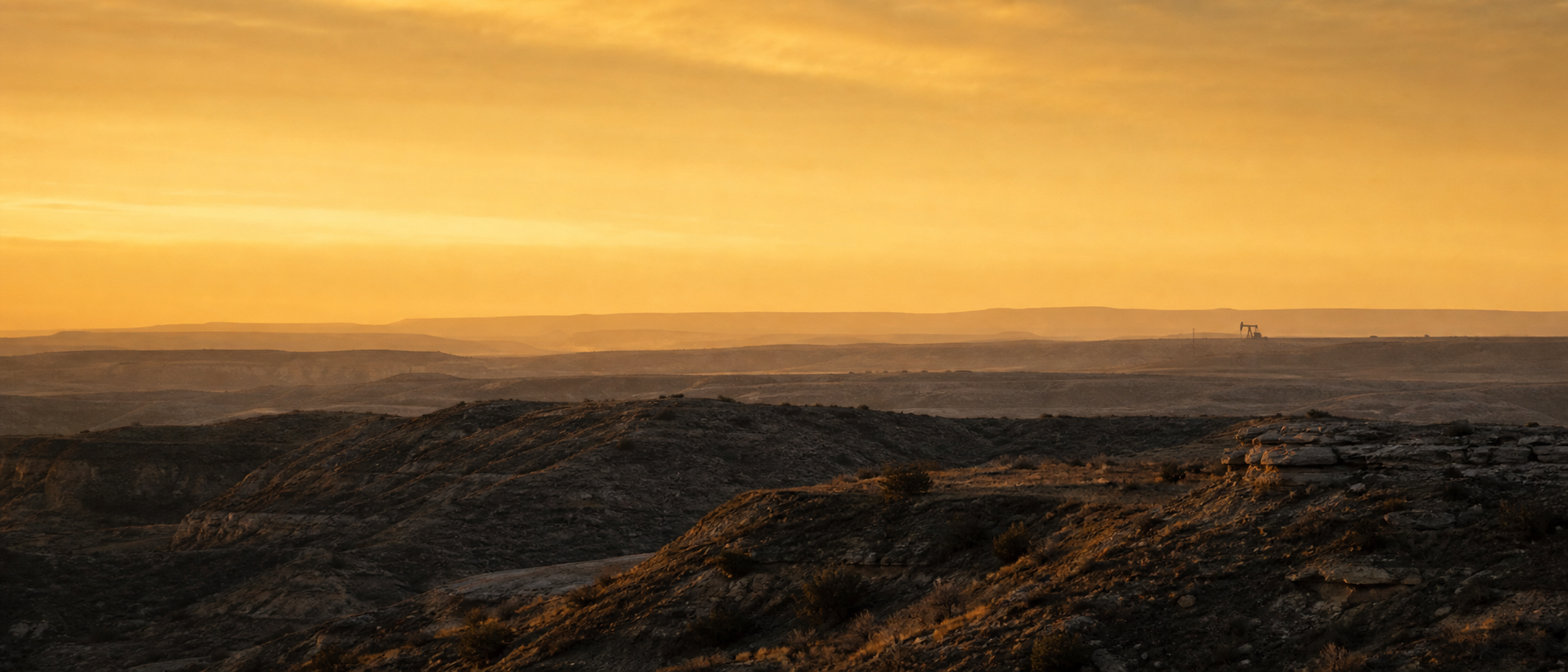 Layered American basin landscape at golden hour, a single distant pumpjack silhouetted on the horizon — the geographies where Peregrine works.